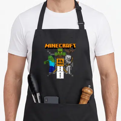 Tablier de cuisine noir avec illustration pixelisée d'ennemis Minecraft Halloween groupés sous le logo orange Minecraft.