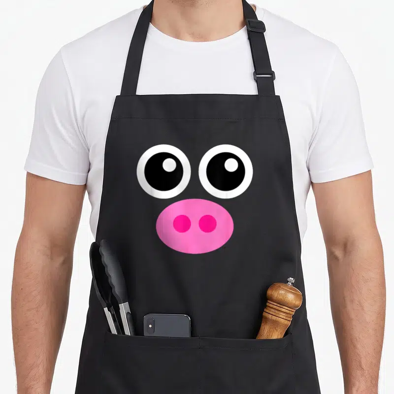 Tablier de cuisine noir avec grands yeux ronds blancs et noirs et un museau de cochon rose vif au centre.
