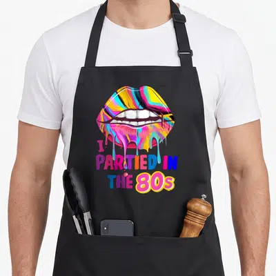 Tablier de cuisine noir avec des lèvres multicolores et le texte I Partied in the 80s en lettres décolorées.