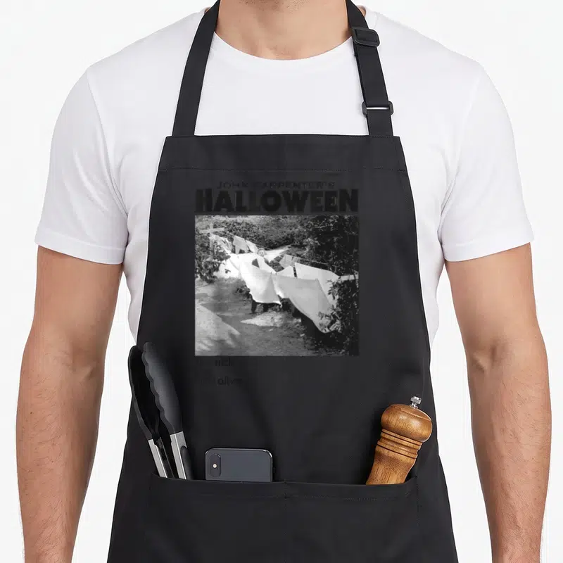 Tablier de cuisine noir avec illustration Michael Myers derrière une corde à linge et texte The trick is to stay alive.