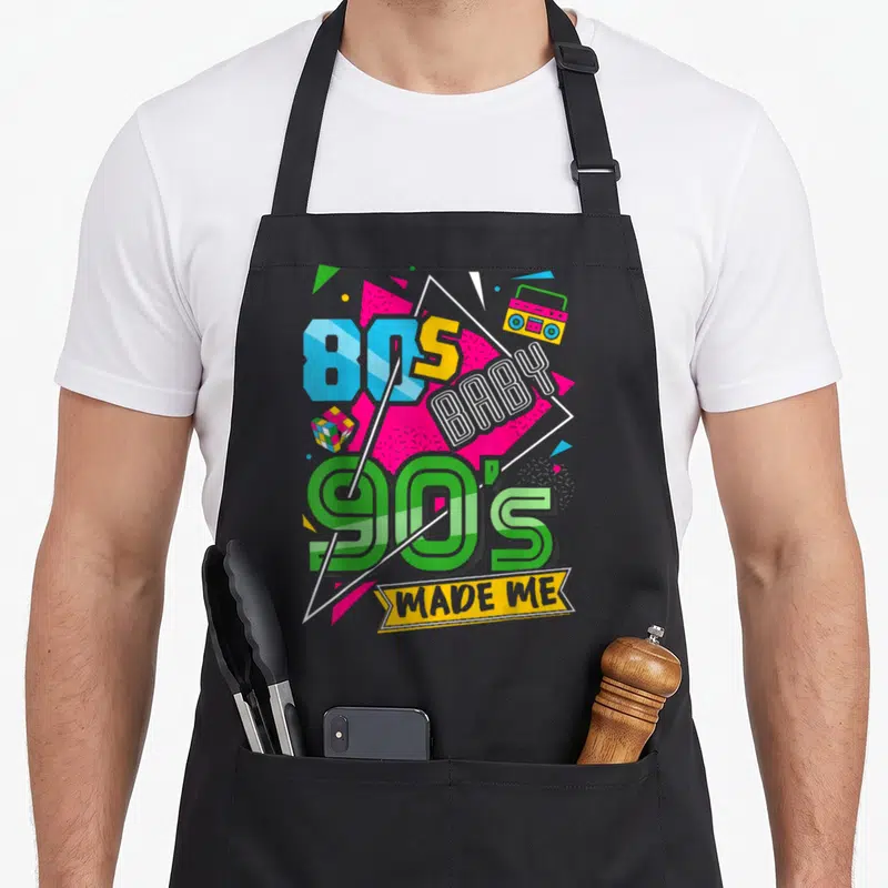 Tablier de cuisine noir avec le texte 80s baby 90s made me et des éléments graphiques colorés sur fond gris.