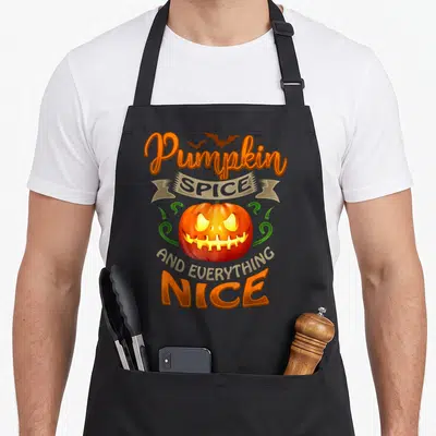 Tablier de cuisine noir avec une citrouille orange aux yeux jaunes et le texte Pumpkin Spice and Everything Nice imprimé en lettres stylisées.