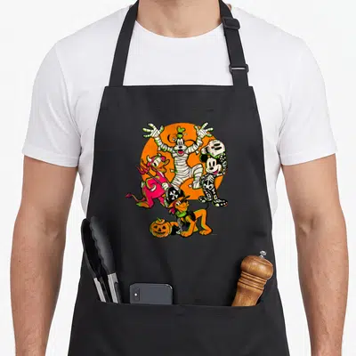 Tablier de cuisine noir avec Mickey squelette, Donald diable, Goofy mummy et Pluto pirate près d'une citrouille.