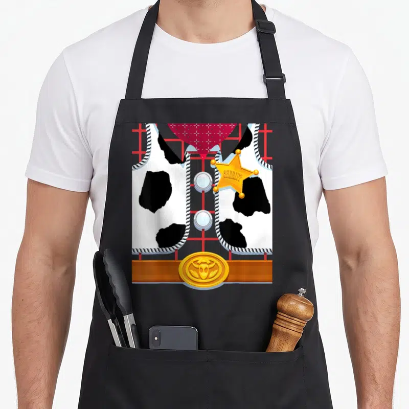 Tablier de cuisine noir avec motif costume de Sheriff Woody, étoile dorée et gilet à pois noirs sur fond quadrillé rouge.