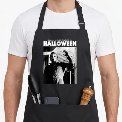 Tablier de cuisine noir avec Michael Myers masqué et texte Halloween en blanc sur fond gris uni.