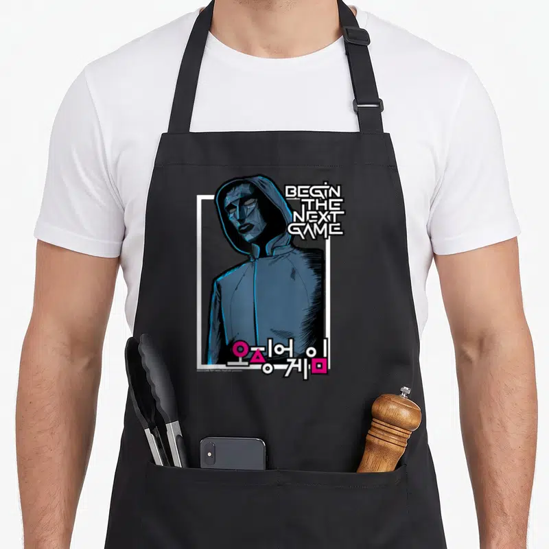 Tablier de cuisine noir avec illustration du Front Man et texte Begin The Next Game en typographie blanche et bleue.