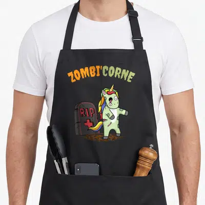 Tablier de cuisine noir avec illustration colorée d'une licorne zombie verte et d'une pierre tombale portant l'inscription RIP.
