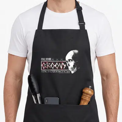 Tablier de cuisine noir avec illustration crâne blanc et texte Groovy Evil Dead en rouge et blanc sur fond uni.