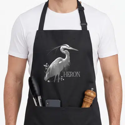 Tablier de cuisine noir avec illustration blanche d'un héron et le mot HERON imprimé en bas à droite.