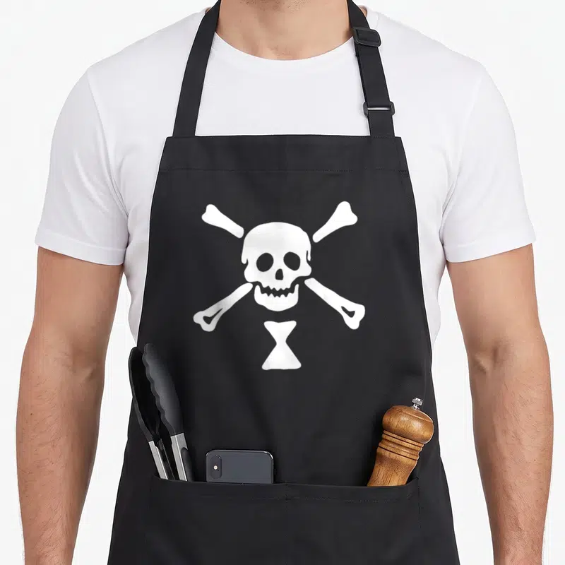 Tablier de cuisine noir avec un drapeau pirate blanc représentant un squelette et des os croisés.