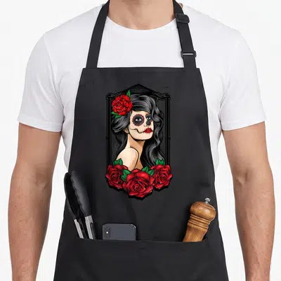 Tablier de cuisine noir avec illustration d'une Catrina aux roses rouges et crâne décoré dans un cadre gothique.