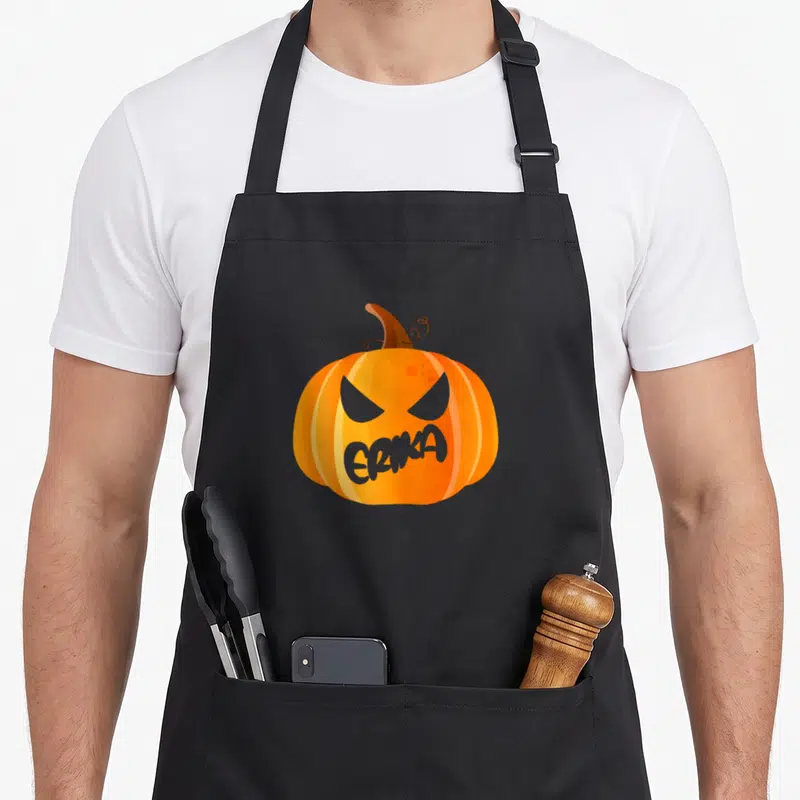 Tablier de cuisine noir avec une grande citrouille orange aux yeux méchants et le nom Erika inscrit sur la bouche.