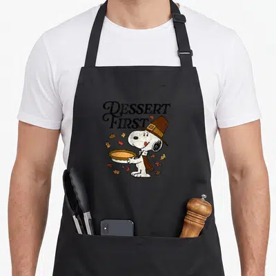 Tablier de cuisine noir avec Snoopy en pèlerin tenant une tarte aux feuilles d'automne autour.