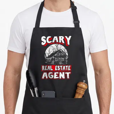 Tablier de cuisine noir avec illustration maison hantée et texte Scary Real Estate Agent en blanc et rouge.