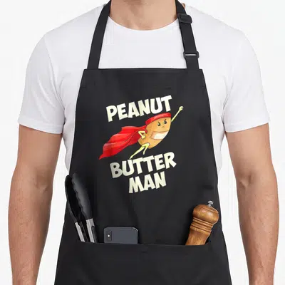 Tablier de cuisine noir avec un super-héros arachide en cape rouge et le texte Peanut Butter Man imprimé en blanc.
