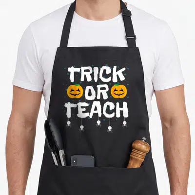 Tablier de cuisine noir avec l'inscription Trick or Teach, deux citrouilles orange et des araignées blanches suspendues.