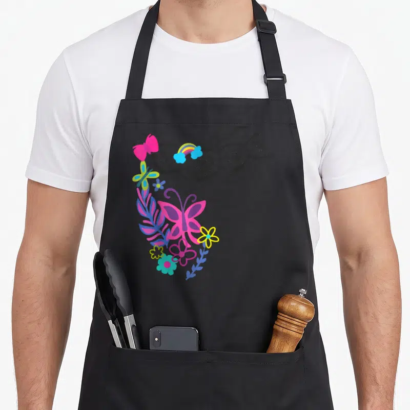 Tablier de cuisine noir avec papillon rose, fleurs multicolores et guirlande noire ornée de petits insectes stylisés dans le coin supérieur gauche.