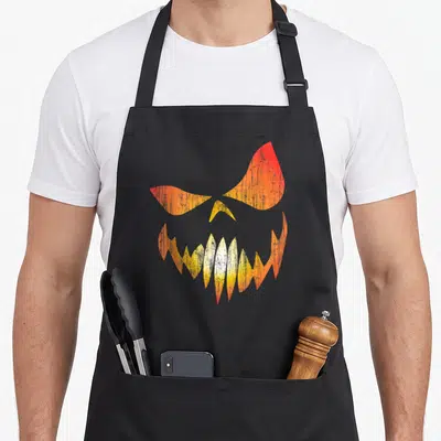 Tablier de cuisine noir avec une citrouille en détresse aux yeux menaçants et des dents pointues dans des tons orange et jaune.