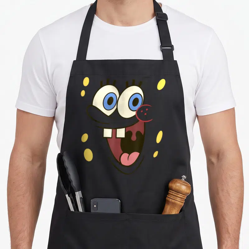 Tablier de cuisine noir avec grand visage cartoon aux yeux bleus, deux dents et bouche ouverte souriante.