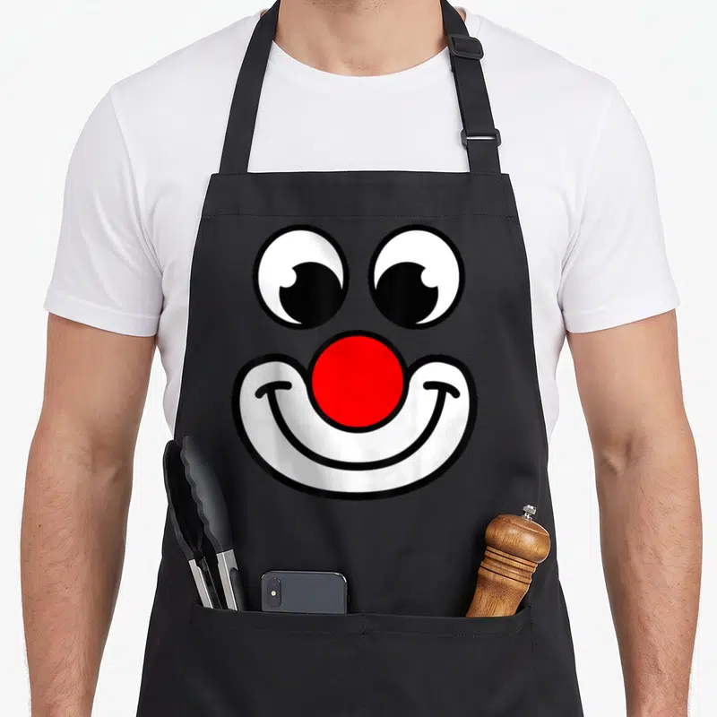 Tablier de cuisine noir orné d'un visage de clown aux yeux ronds, nez rouge et sourire blanc stylisé.
