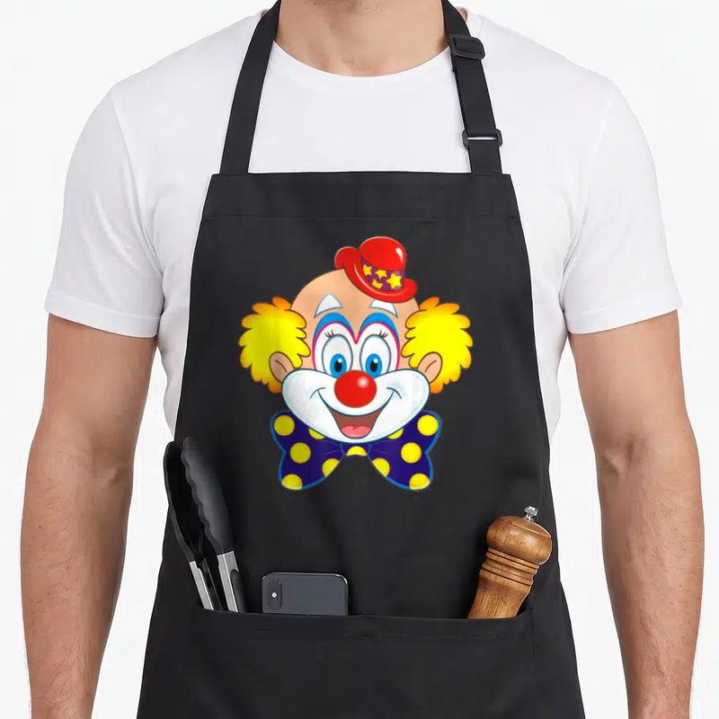 Tablier de cuisine noir avec une illustration de tête de clown souriant au chapeau rouge et nœud papillon bleu à pois jaunes.
