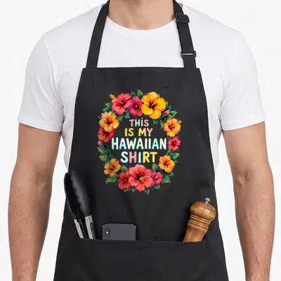 Tablier de cuisine noir orné d'un cercle de fleurs hibiscus et du texte This Is My Hawaiian Shirt au centre.