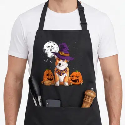Tablier de cuisine noir avec illustration d'un corgi en chapeau de sorcière entouré de citrouilles effrayantes sous une pleine lune.