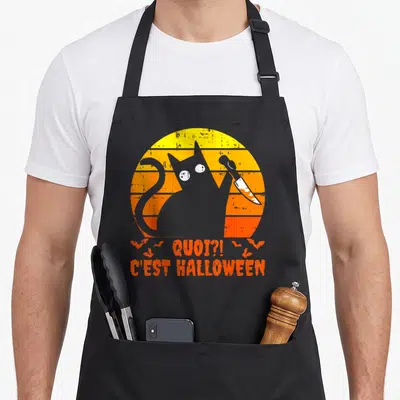 Tablier de cuisine noir avec un chat gris tenant un couteau et le texte Quoi ?. C'est Halloween sur fond orange.