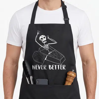 Tablier de cuisine noir avec un squelette blanc sortant d'un coffre et le texte Never Better imprimé en dessous.