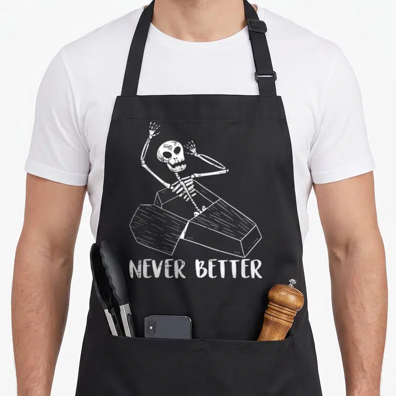 Tablier de cuisine noir avec un squelette blanc sortant d'un coffre et le texte Never Better imprimé en dessous.