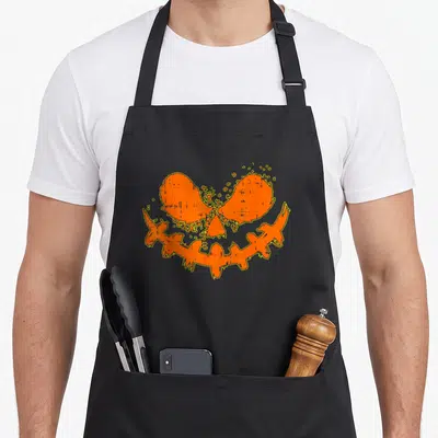 Tablier de cuisine noir avec un visage de pumpkin orange stylisé et du texte humoristique en haut.
