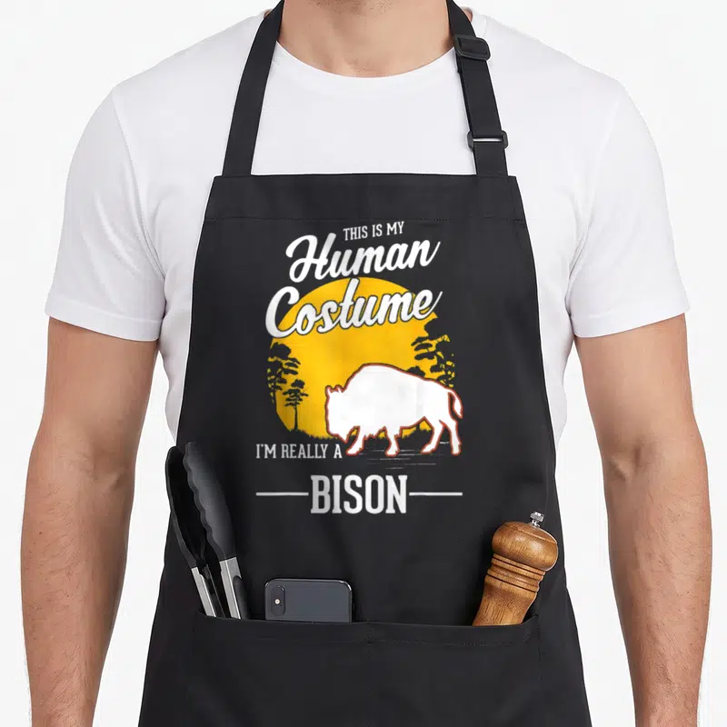 Tablier de cuisine noir avec illustration d'un bison blanc et texte humoristique sur fond orange.
