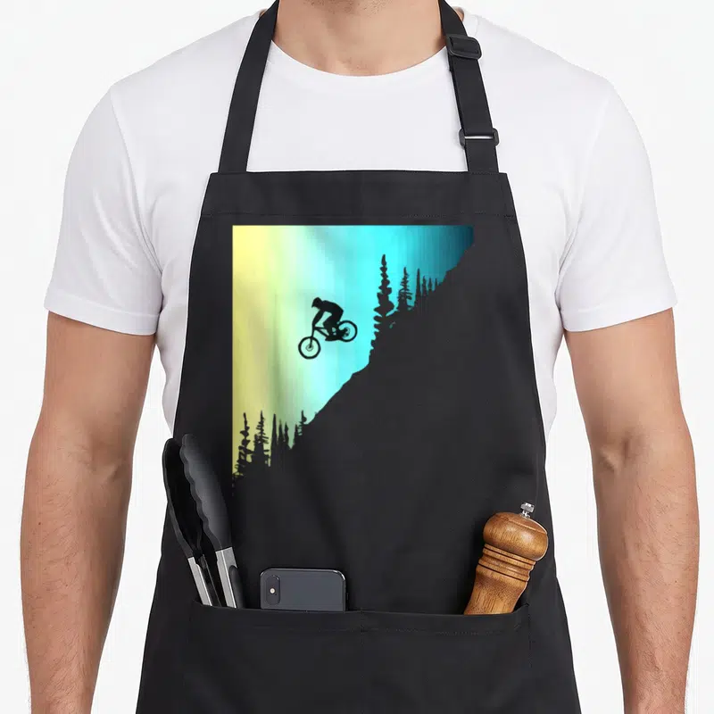 Tablier de cuisine noir avec silhouette cycliste en tenue de descente sur fond dégradé jaune et bleu.