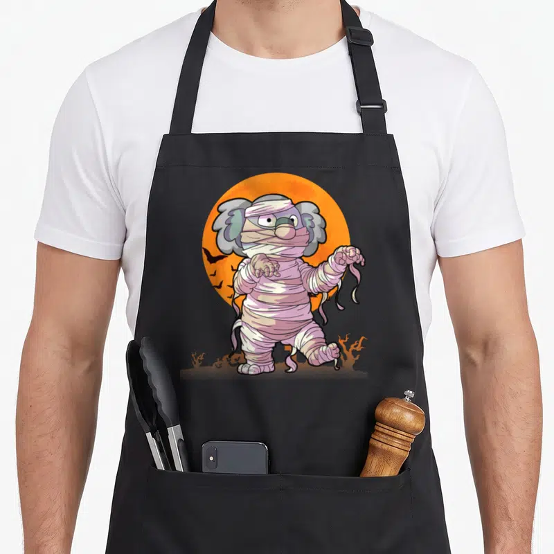 Tablier de cuisine noir avec illustration d'un koala momie aux bandages roses devant une lune orange.