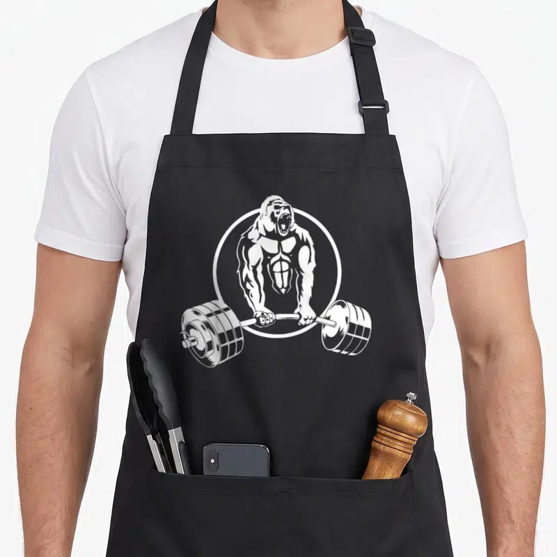Tablier de cuisine noir avec illustration blanche d'un gorille musclé tenant une barre de poids lourde dans un cercle.