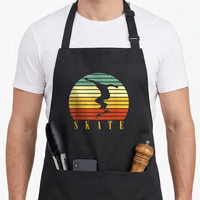 Tablier de cuisine noir avec silhouette de skateur sur fond de soleil rayé vert jaune orange et texte SKATE doré.