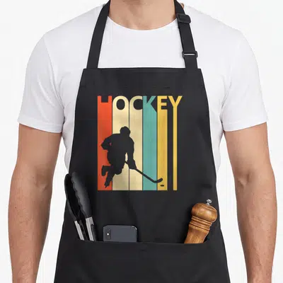 Tablier de cuisine noir avec silhouette de joueur de hockey sur glace devant des bandes de couleur et le mot HOCKEY.