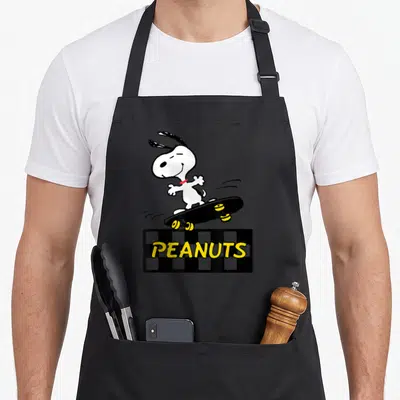 Tablier de cuisine noir avec Snoopy sur planche à roulettes et logo Peanuts en lettres jaunes sur fond damier.