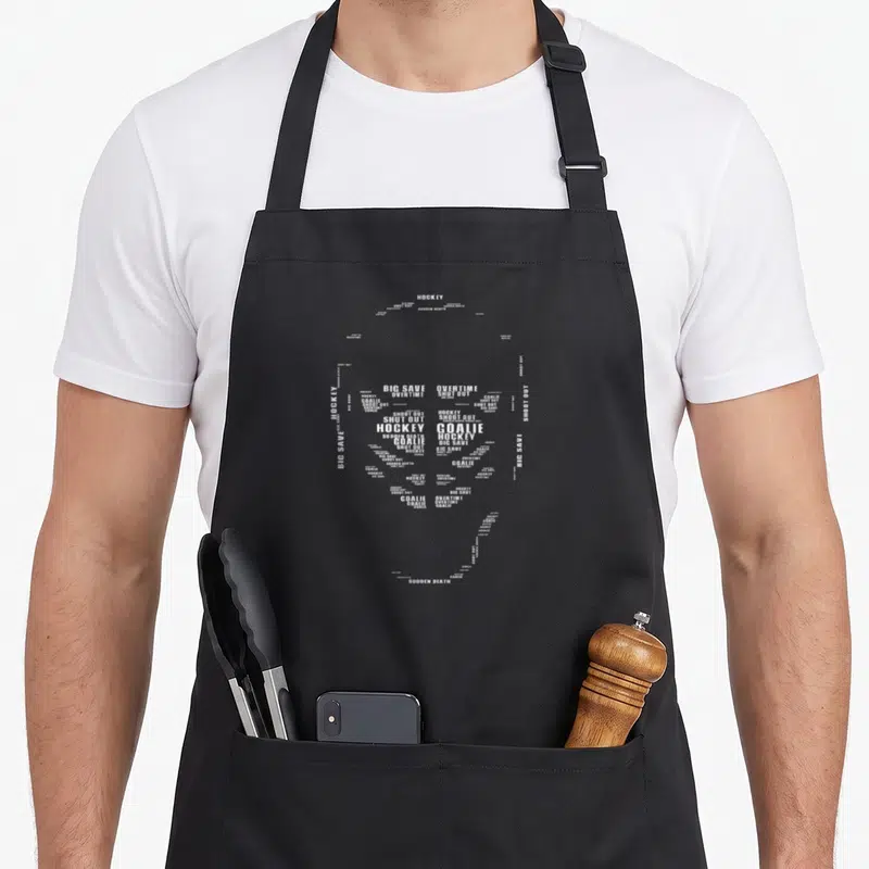 Tablier de cuisine noir avec un masque de gardien de hockey composé de mots blancs comme Goalie et Big Save.