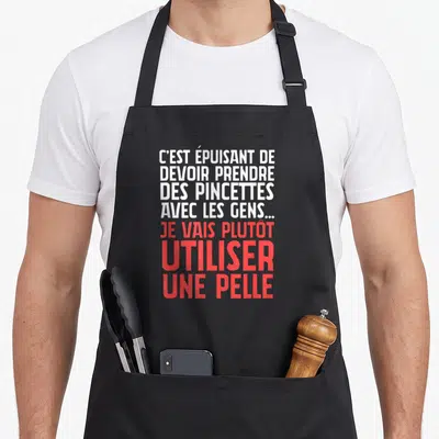 Tablier de cuisine noir avec citation blanche et rouge sur l'humour des pincettes et de la pelle pour le barbecue.