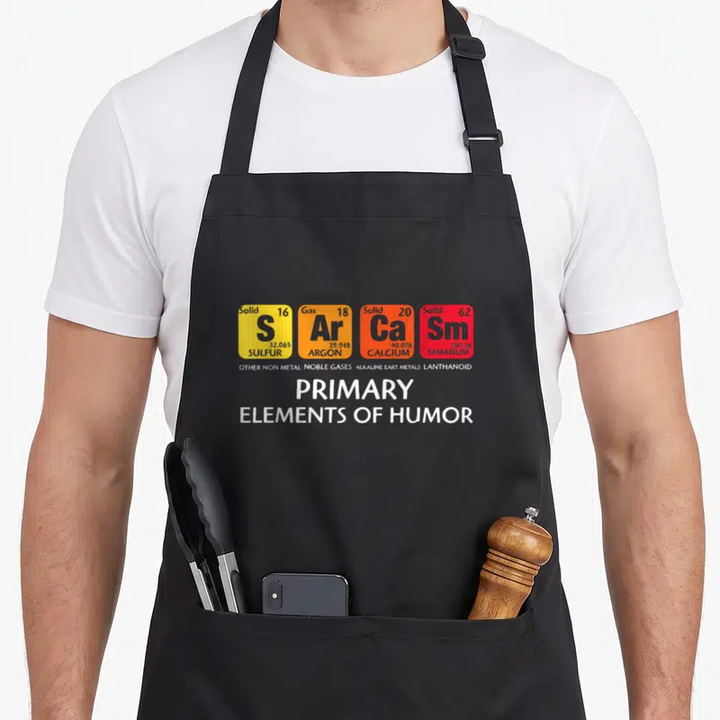 Tablier de cuisine noir avec quatre cases colorées imitant des éléments chimiques et le texte PRIMARY ELEMENTS OF HUMOR en blanc.