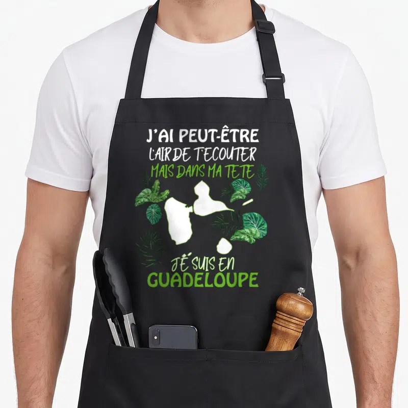 Tablier de cuisine noir avec carte blanche de la Guadeloupe entourée de feuilles vertes et texte humoristique sur le fond.