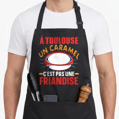 Tablier de cuisine noir avec un ballon de rugby et le texte un caramel c'est pas une friandise en lettres colorées.