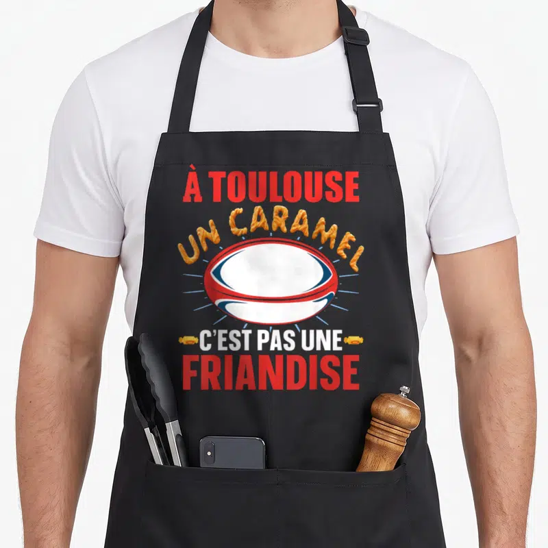 Tablier de cuisine noir avec un ballon de rugby et le texte un caramel c'est pas une friandise en lettres colorées.