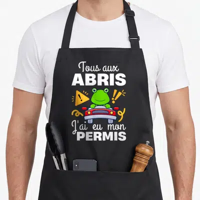 Tablier de cuisine noir avec une grenouille verte au volant d'une voiture rouge et le texte Tous aux abris j'ai eu mon permis.