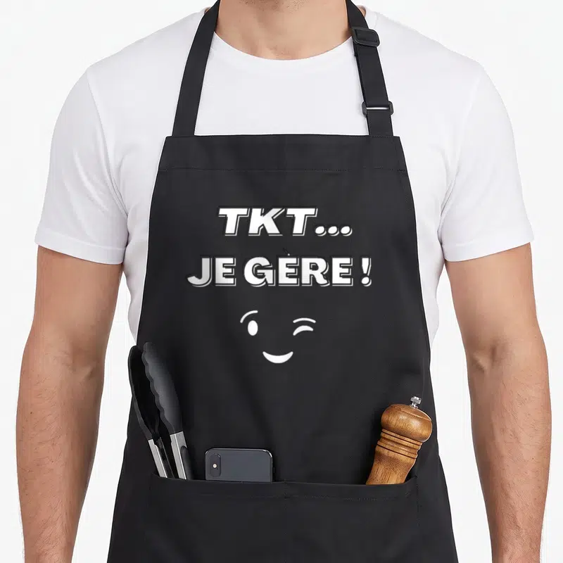 Tablier de cuisine noir avec l'inscription TKT JE GERE et un visage souriant qui fait un clin d'oeil en blanc.