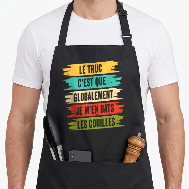 Tablier de cuisine noir avec le texte « Le truc c'est que globalement je m'en bats les couilles » sur fond de traits colorés.