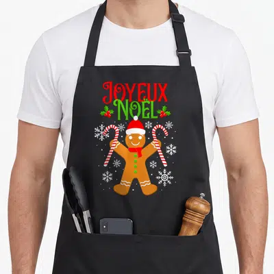 Tablier de cuisine noir avec un biscuit de Noël tenant des cannes à sucre et le texte Joyeux Noël en lettres rouges et vertes.