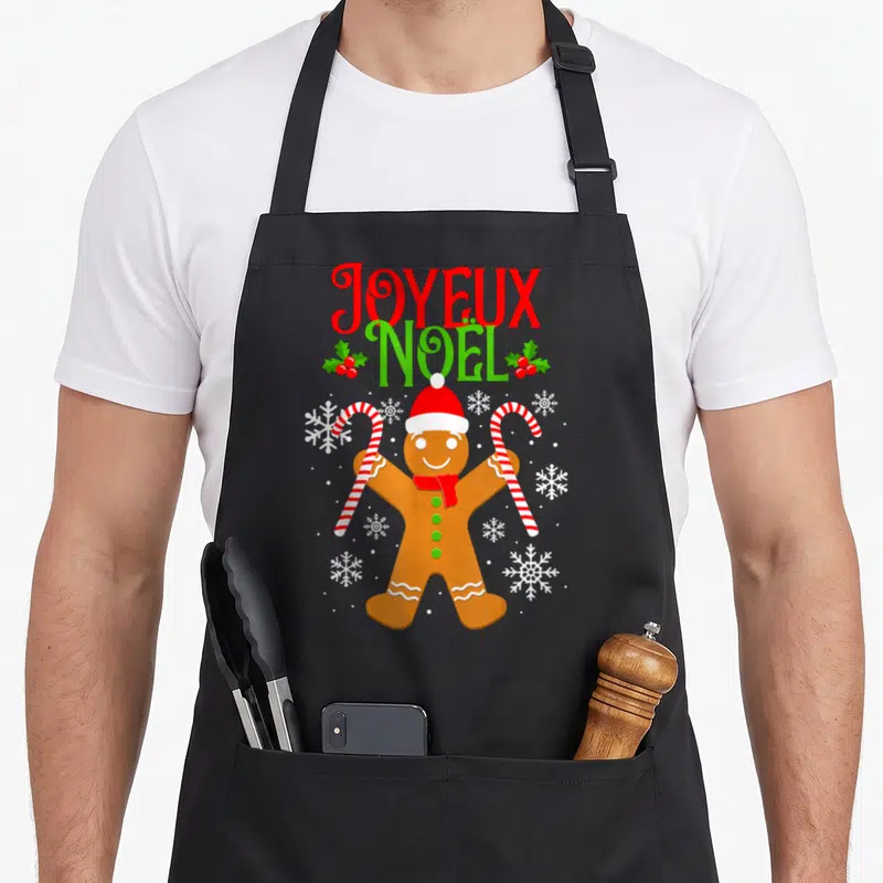 Tablier de cuisine noir avec un biscuit de Noël tenant des cannes à sucre et le texte Joyeux Noël en lettres rouges et vertes.