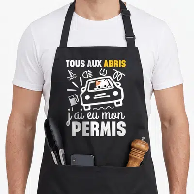 Tablier de cuisine noir avec illustration d'un chat au volant et texte tous aux abris j'ai eu mon permis en blanc et orange.